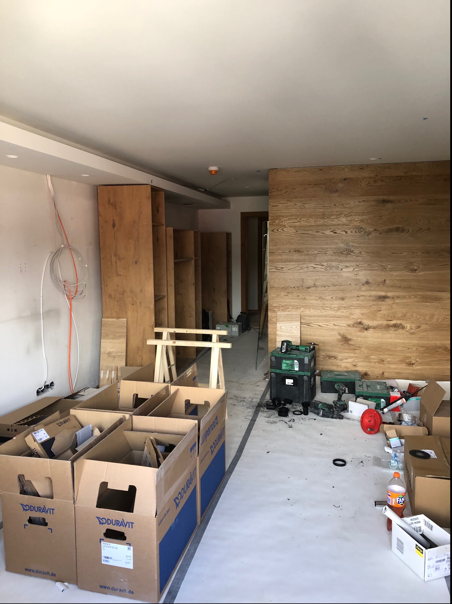 Wnętrze w trakcie prac wykończeniowych: montaż drewnianej zabudowy, widoczne kartony z logo Duravit, narzędzia i kable elektryczne na ścianie.