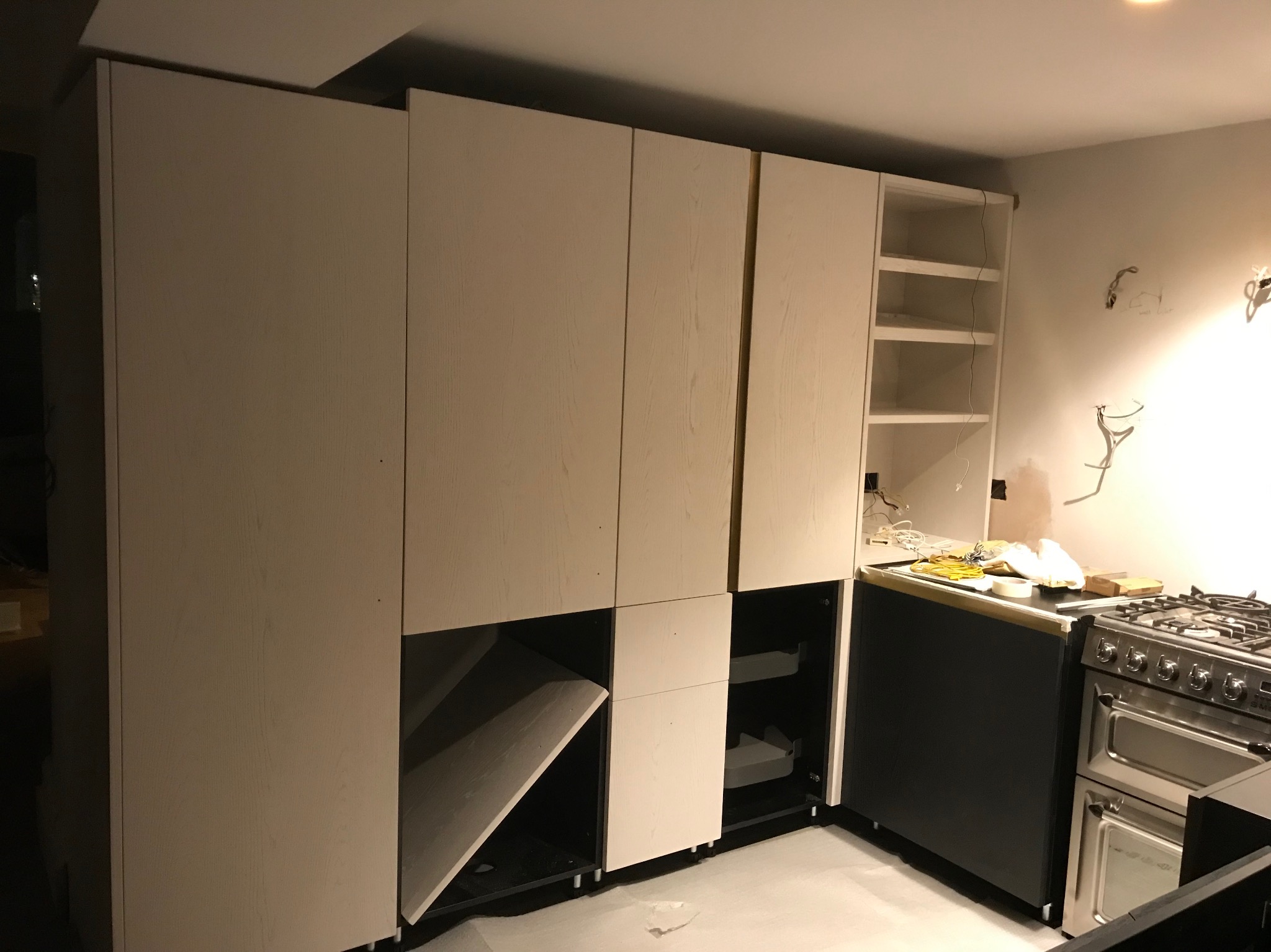 Montaż szafy wnękowej z jasnego drewna w kuchni, widoczne otwarte półki i szuflady, częściowo zamontowany front, obok kuchenka gazowa.