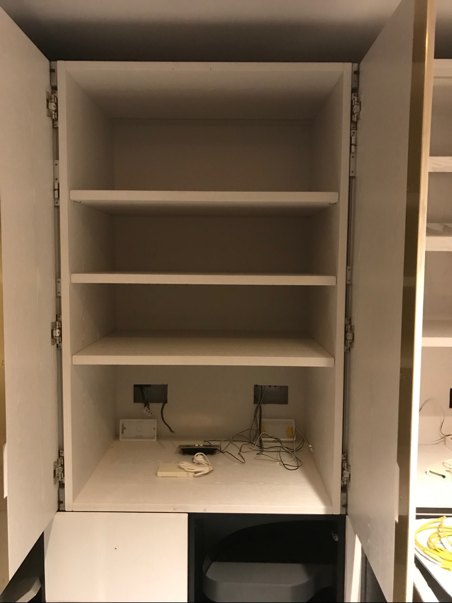 Wnętrze białej szafy wnękowej z otwartymi drzwiami, widoczne trzy półki, okablowanie elektryczne w dolnej części i elementy montażowe.