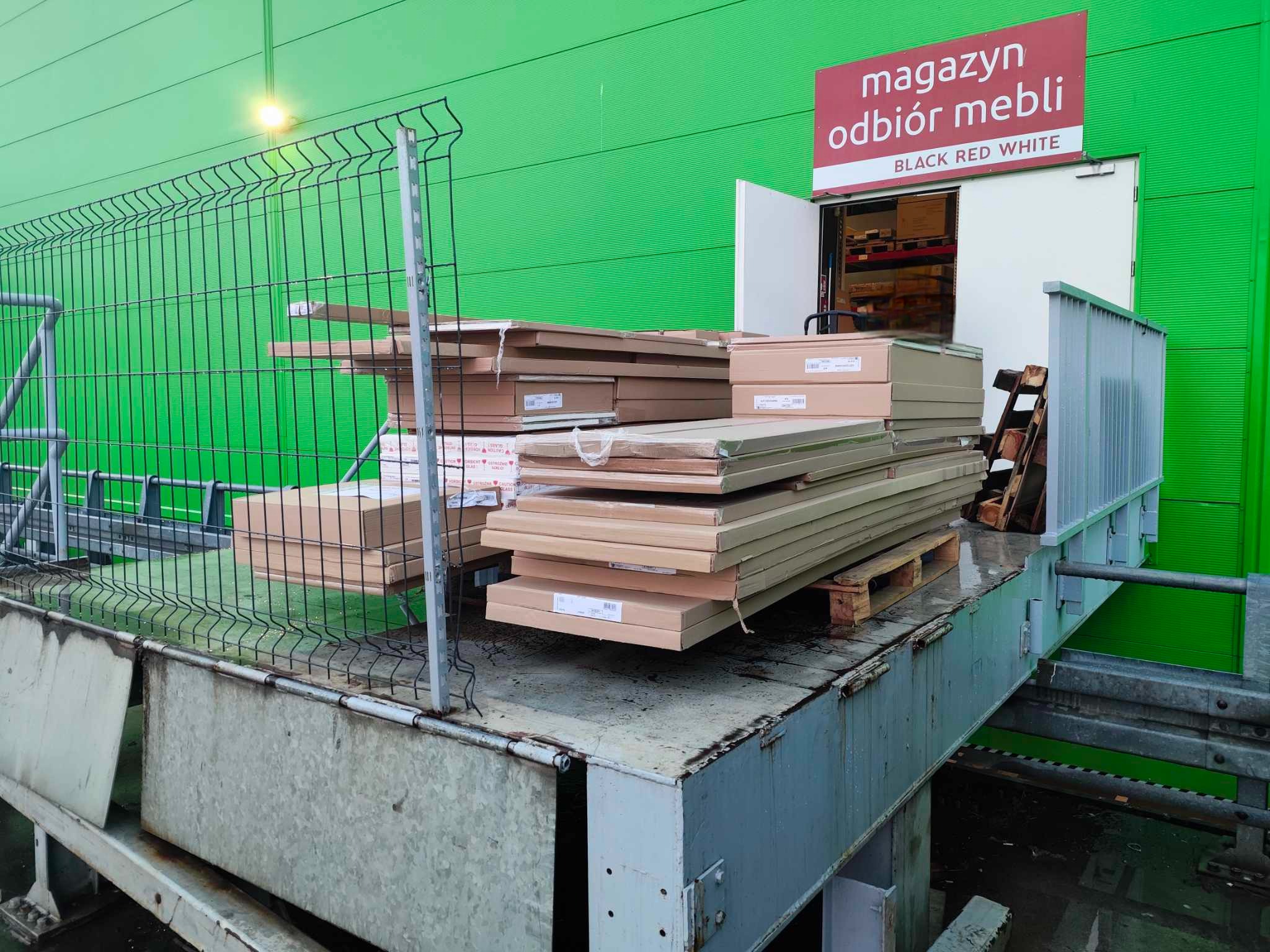 Paleta z zapakowanymi elementami meblowymi przed magazynem odbioru mebli Black Red White, widoczna zielona ściana magazynu i metalowe ogrodzenie.
