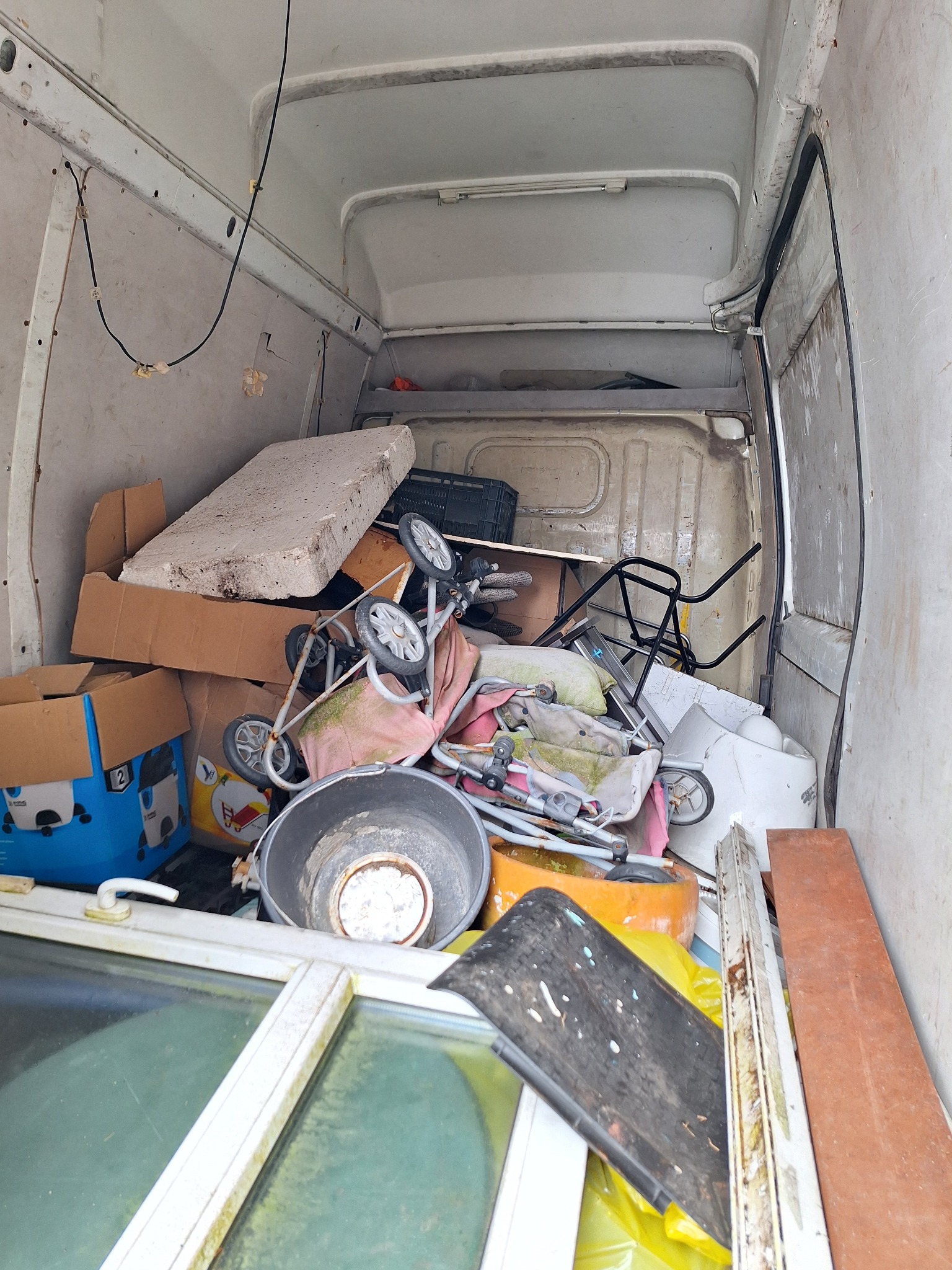 Wnętrze furgonetki wypełnionej przedmiotami do przewiezienia, w tym okno w białej ramie, dziecięcy wózek, kartony i wiadro.