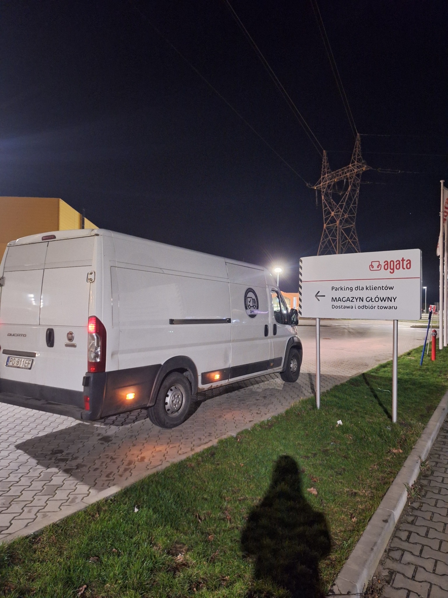 Biały bus dostawczy marki Ducato zaparkowany nocą obok znaku informacyjnego 'Agata: Parking dla klientów, Magazyn Główny, Dostawa i odbiór towaru', z widoczną w tle linią wysokiego napięcia.
