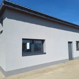 Jasnoszary budynek z nową elewacją, ciemnoszare okna i drzwi, widoczna faktura tynku baranka, szara podmurówka, kostka brukowa na podjeździe, błękitne niebo.