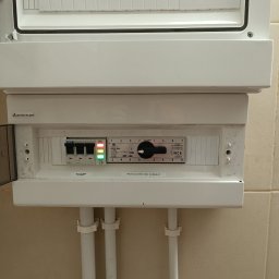 Dam-Pol - Otwarta skrzynka rozdzielcza Elektro-Plast z widocznymi bezpiecznikami Eaton, przełącznikiem obrotowym i diodami LED, zamontowana na ścianie z beżowych płytek, z trzema białymi rurami...