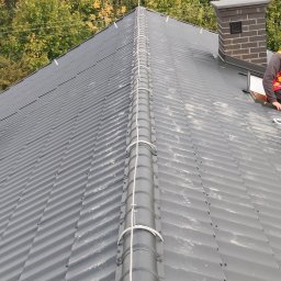 Dam-Pol - Szary blaszany dach z zamontowanym systemem przewodów odprowadzających ładunki elektryczne, biegnących wzdłuż kalenicy, zakończonych pionowym masztem, z kominem w tle i fragmentem sylwetki...