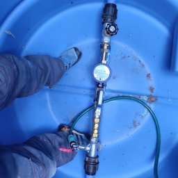 Dam-Pol - Wnętrze niebieskiej studni wodomierzowej z zamontowanym licznikiem, zaworami i rurami z tworzywa sztucznego; widoczne fragmenty ubrania i butów osoby instalującej.