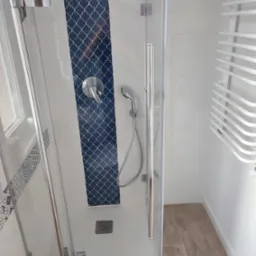 Nowoczesna kabina prysznicowa z chromowanymi elementami, zdobiona pionowym pasem płytek w kolorze granatowym z białym wzorem marokańskim, białe płytki na ścianach, drewnopodobna podłoga i biały...