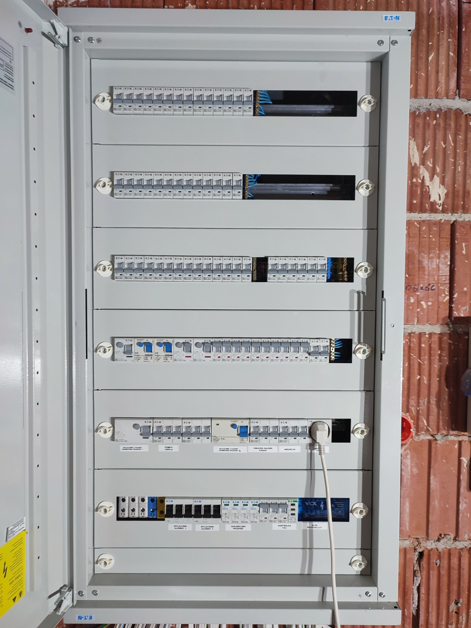 Otwarta szara rozdzielnica elektryczna marki Eaton zamontowana na czerwonej ceglanej ścianie, widoczne zabezpieczenia, wyłączniki i podłączony przewód z wtyczką.