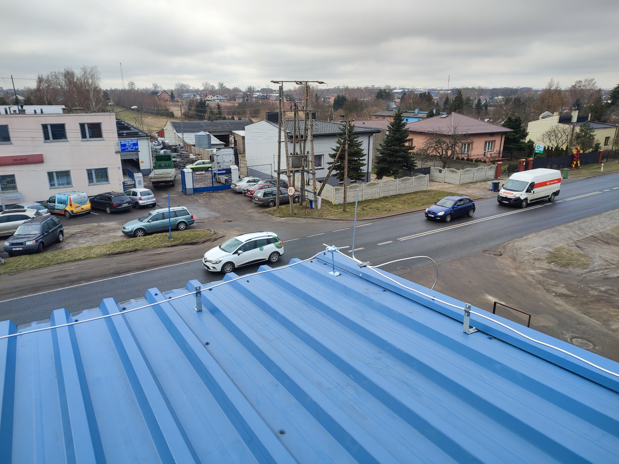 Błękitny blaszany dach z zamontowaną instalacją odgromową, widok z góry na ulicę z przejeżdżającymi samochodami i zabudowę miejską w tle.