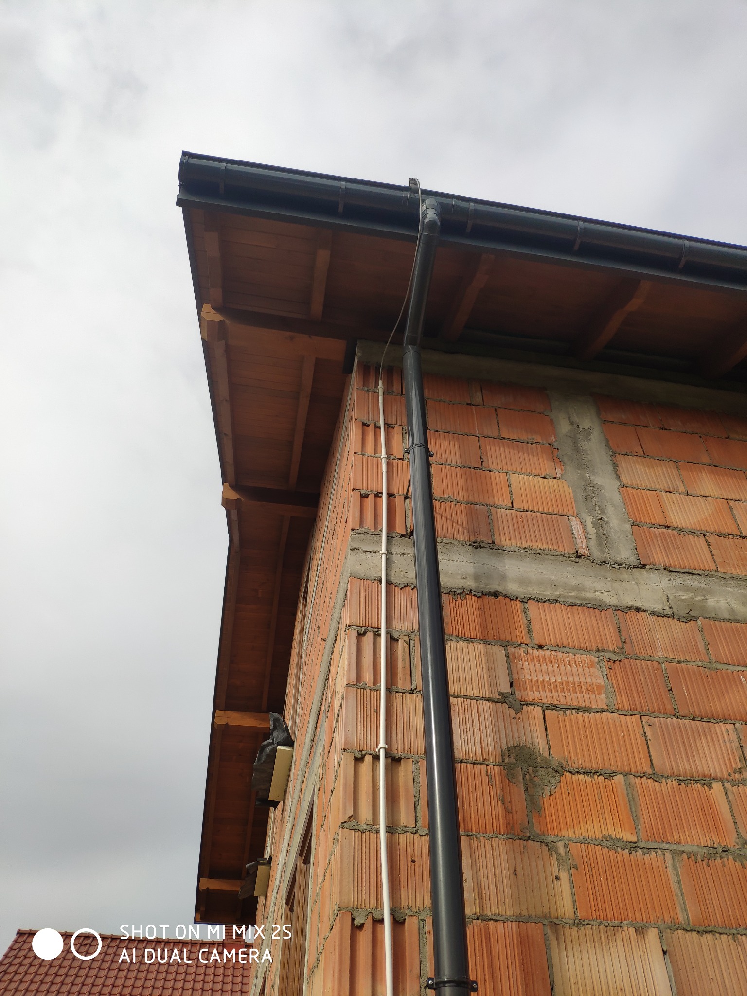 Rzut z dołu na róg budynku w trakcie budowy z zamontowanym systemem odprowadzania wody deszczowej; widoczny surowy mur z cegły dziurawki, drewniane elementy dachu i rynna spustowa.