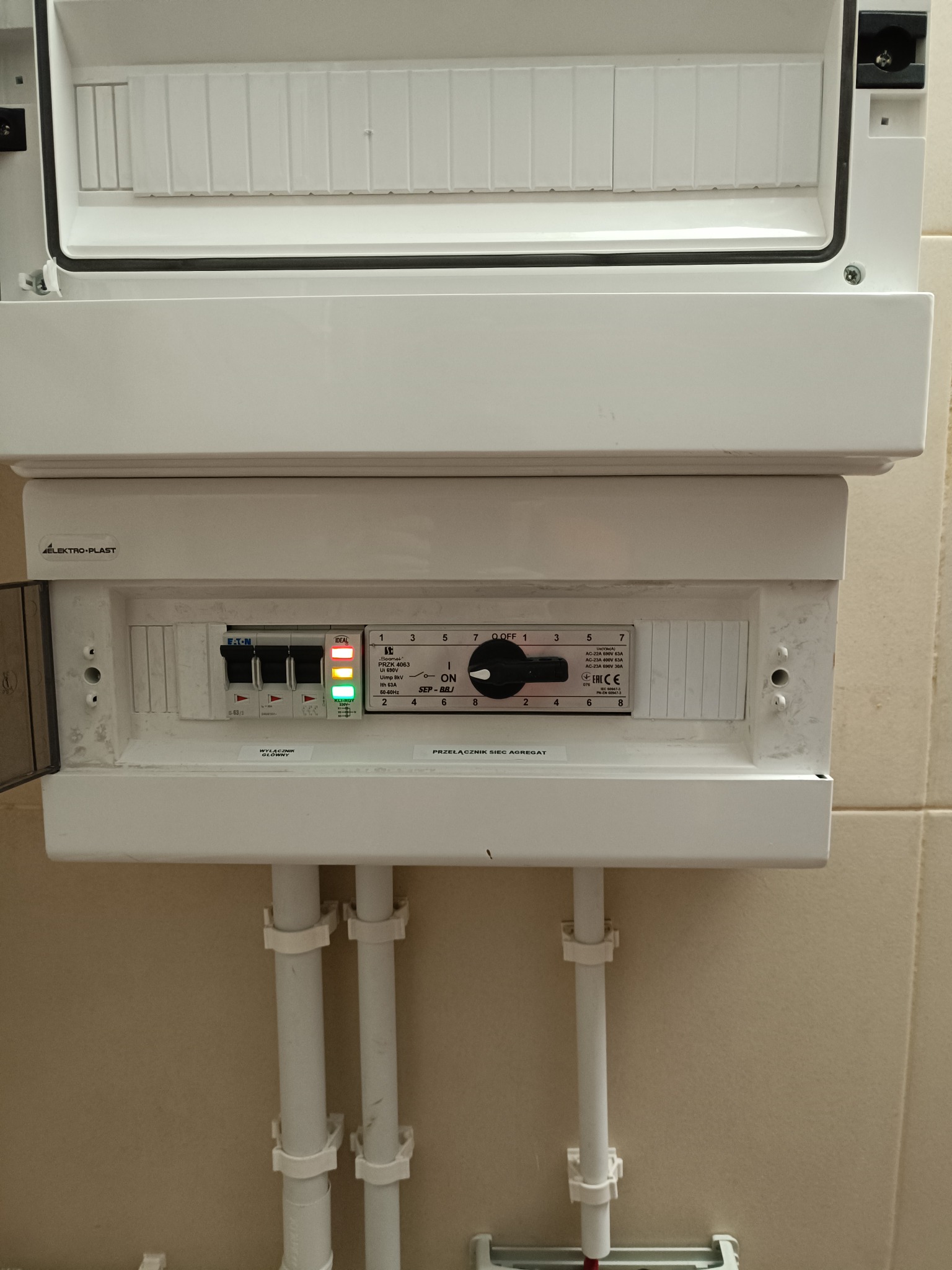 Otwarta skrzynka rozdzielcza Elektro-Plast z widocznymi bezpiecznikami Eaton, przełącznikiem obrotowym i diodami LED, zamontowana na ścianie z beżowych płytek, z trzema białymi rurami...