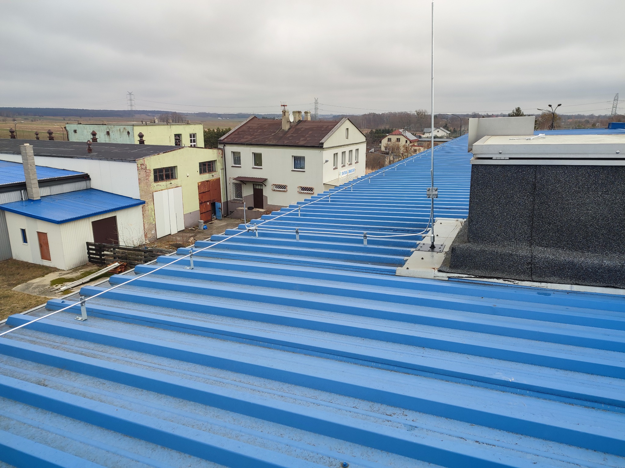 Błękitny, blaszany dach przemysłowy z zainstalowanym systemem ochrony odgromowej: maszt odgromowy na szczycie budynku, linka poprowadzona wzdłuż dachu za pomocą uchwytów. W tle budynki gospodarcze...