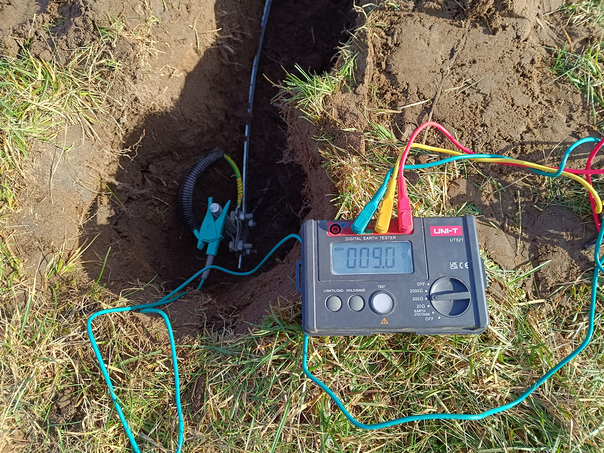 Pomiar rezystancji uziemienia za pomocą miernika UNI-T UT521, z widocznymi kolorowymi przewodami i złączem uziemiającym w wykopie ziemnym.
