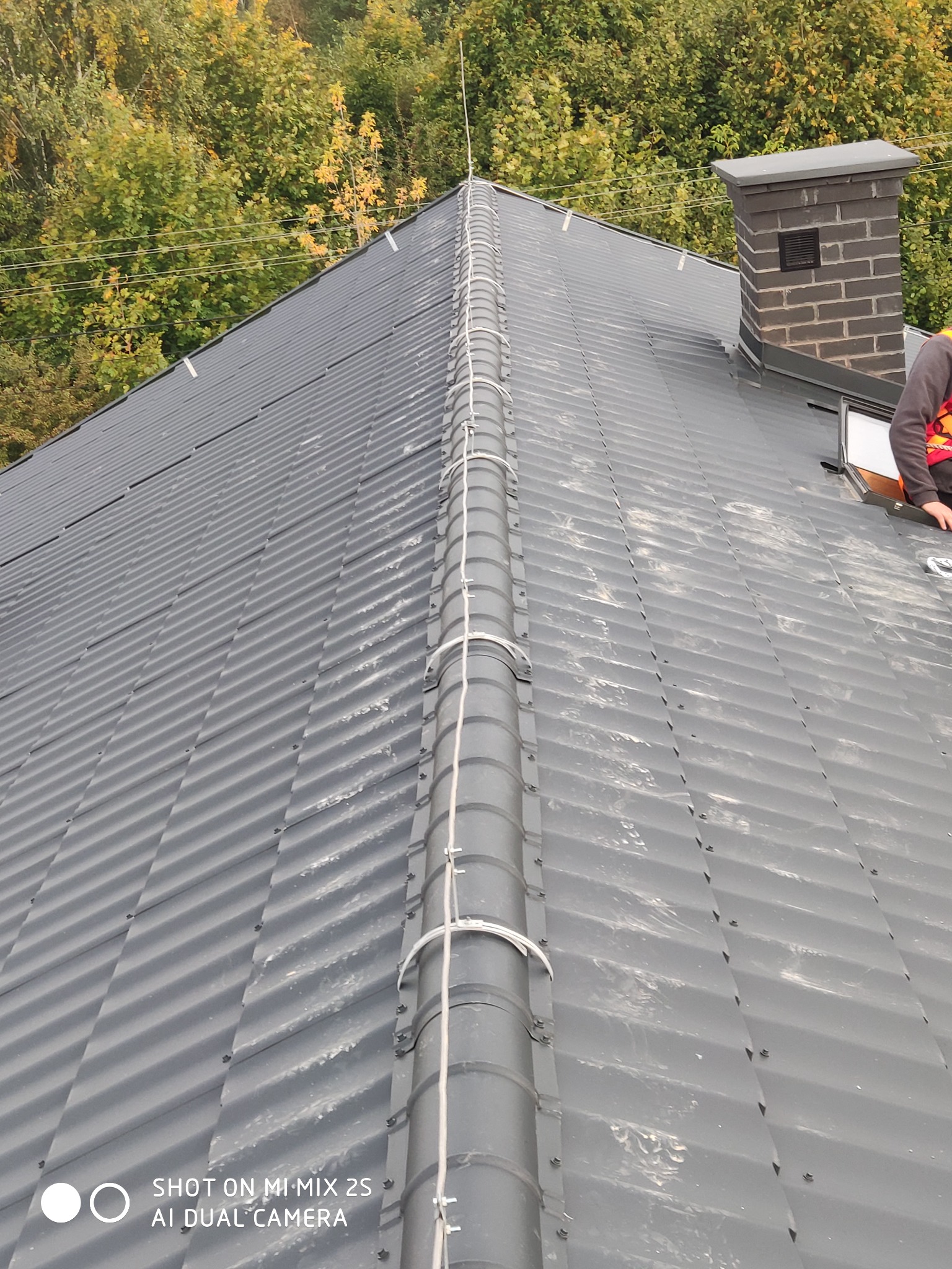 Szary blaszany dach z zamontowanym systemem przewodów odprowadzających ładunki elektryczne, biegnących wzdłuż kalenicy, zakończonych pionowym masztem, z kominem w tle i fragmentem sylwetki...