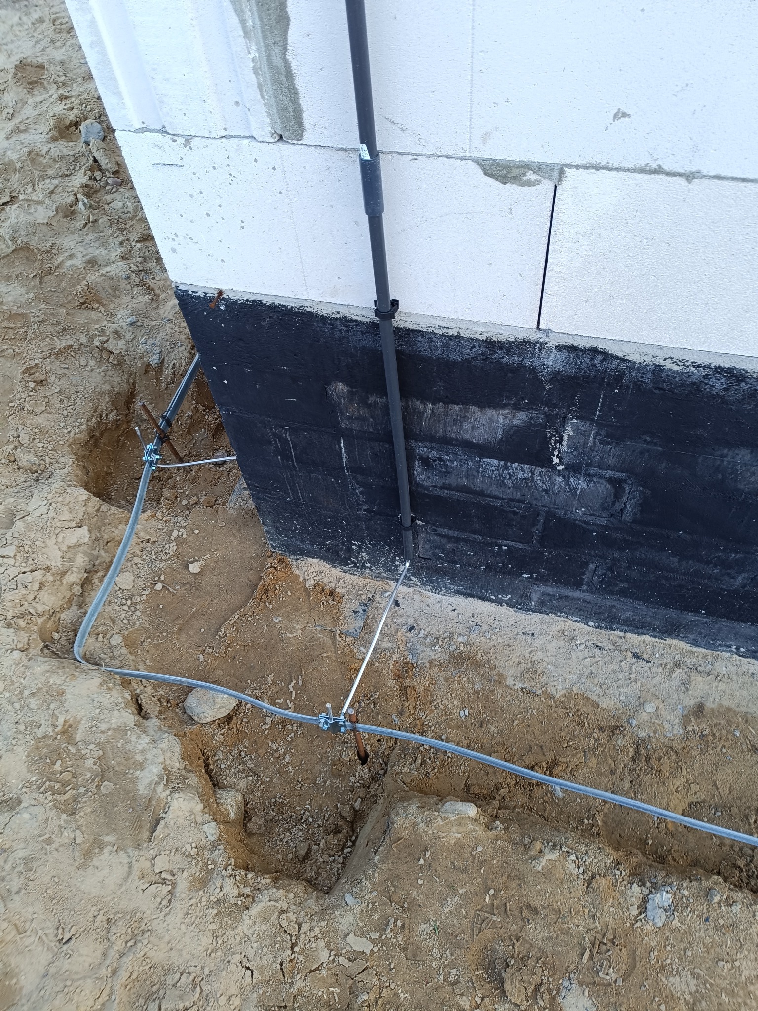 Uziemienie budynku w trakcie instalacji: widoczne przewody uziemiające prowadzone w wykopie wzdłuż fundamentu zabezpieczonego czarną izolacją, połączone z pionowym przewodem mocowanym do ściany...