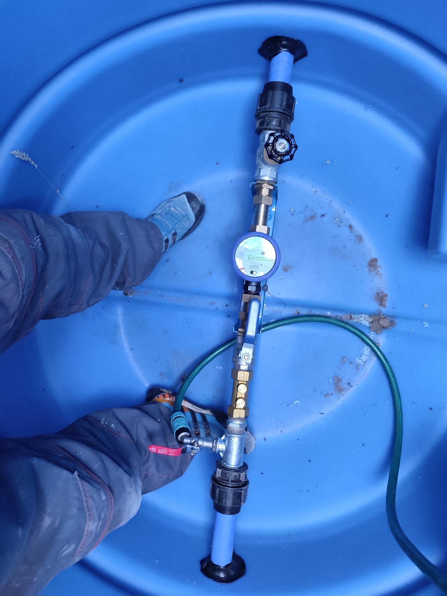 Wnętrze niebieskiej studni wodomierzowej z zamontowanym licznikiem, zaworami i rurami z tworzywa sztucznego; widoczne fragmenty ubrania i butów osoby instalującej.