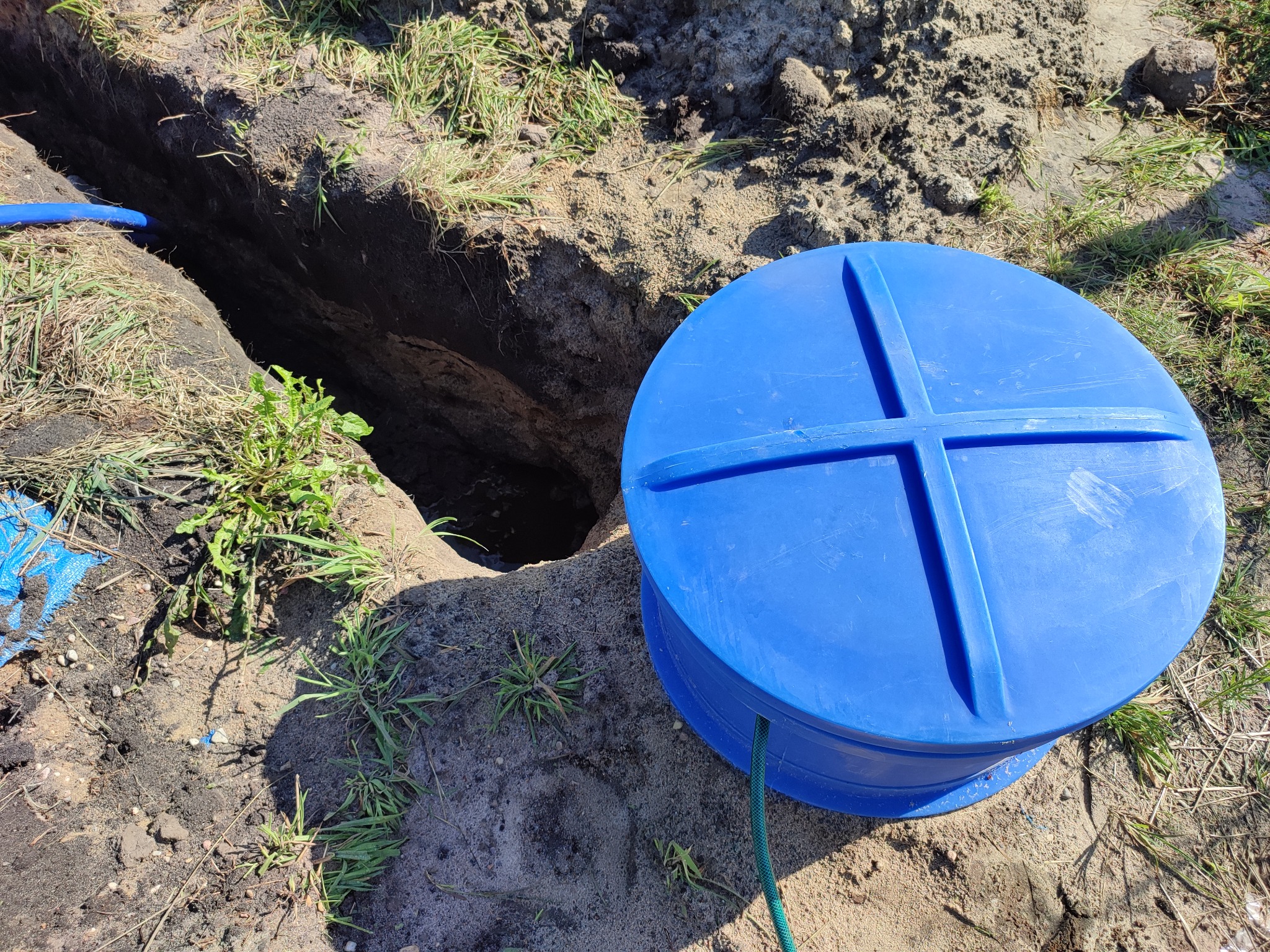 Błękitna pokrywa studni wodomierzowej z charakterystycznym krzyżowym wzmocnieniem, umieszczona obok wykopu w ziemi w Łowiczu, z widocznym fragmentem niebieskiego przewodu w rowie.