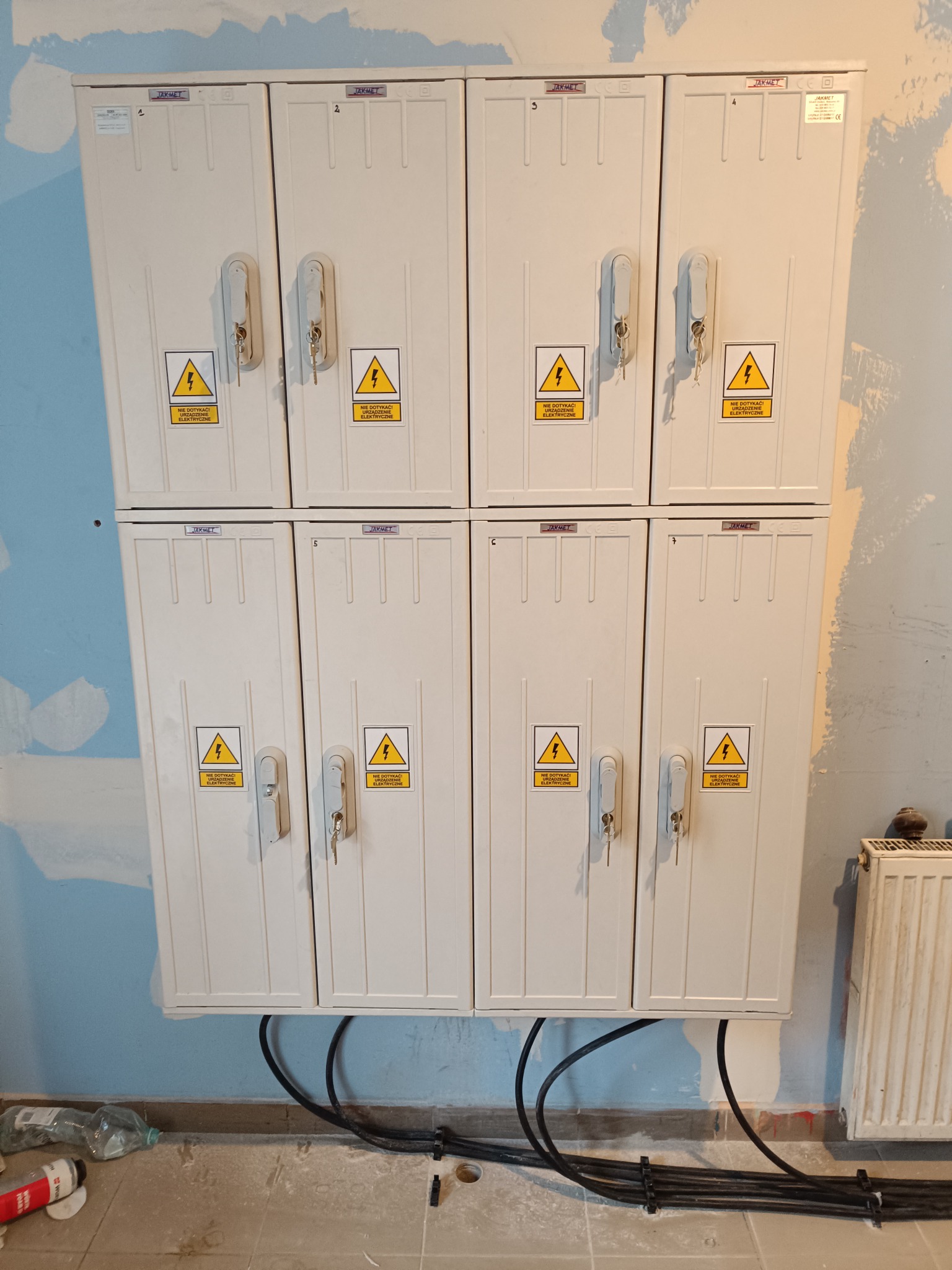 Zestaw ośmiu skrzynek licznikowych Jakmet z kluczykami, umieszczonych w dwóch rzędach po cztery, z żółtymi naklejkami ostrzegawczymi, zamontowanych na ścianie w trakcie remontu, z widocznymi...