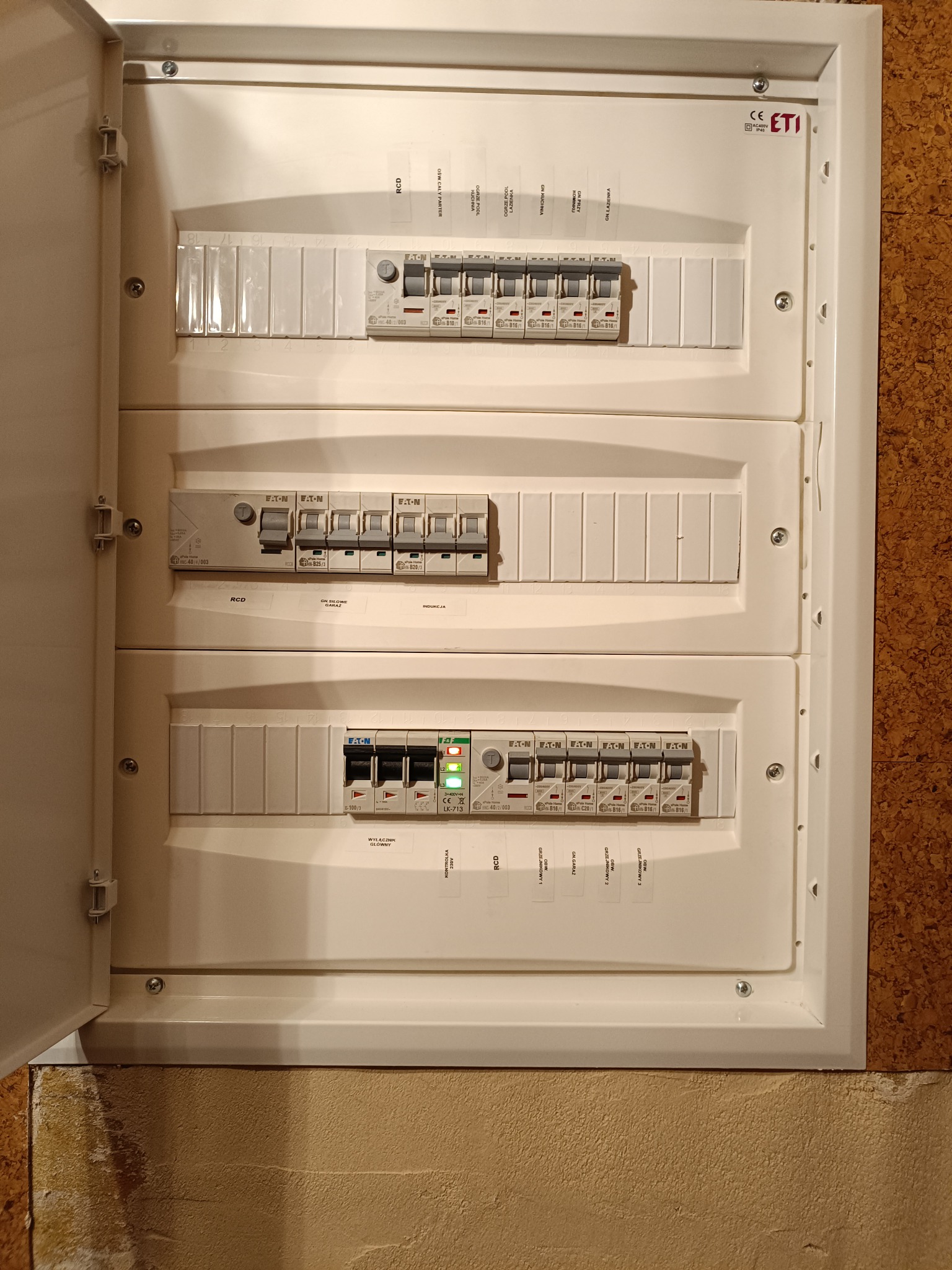 Otwarta skrzynka rozdzielcza z trzema rzędami wyłączników Eaton, widoczne opisy obwodów i kontrolka 230V, zamontowana na ścianie o nierównej fakturze.