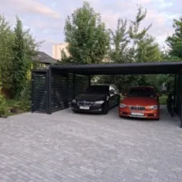 Dwa samochody zaparkowane pod nowoczesną, czarną, metalową wiatą garażową z ażurowymi ścianami bocznymi, na posesji z kostki brukowej, otoczonej zielenią.