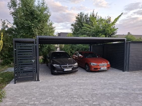 Dwa samochody, czarny i pomarańczowy, zaparkowane pod nowoczesną, metalową wiatą samochodową z bocznymi panelami i ażurową furtką, na posesji wyłożonej kostką brukową.