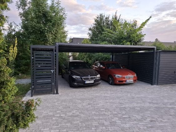 Dwa samochody zaparkowane pod nowoczesną, metalową wiatą garażową z ażurowymi panelami bocznymi, obok garażu blaszanego, na posesji z kostki brukowej.