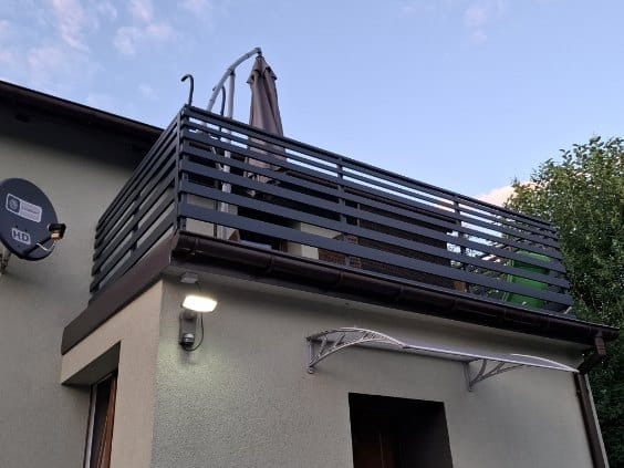 Czarna, pozioma balustrada na balkonie zadaszonym brązowym dachem, widok z dołu, antena satelitarna na ścianie, szara elewacja budynku, markiza nad wejściem.