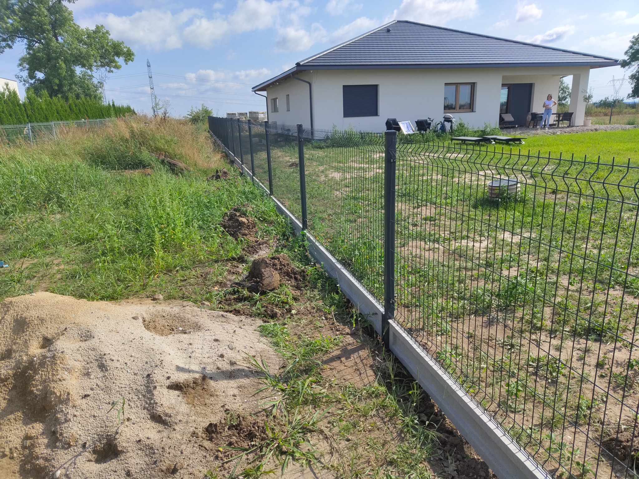 Montaż ogrodzenia panelowego z podmurówką betonową, widoczna sterta piasku i trawa, w tle biały dom z szarym dachem i osoba na tarasie.