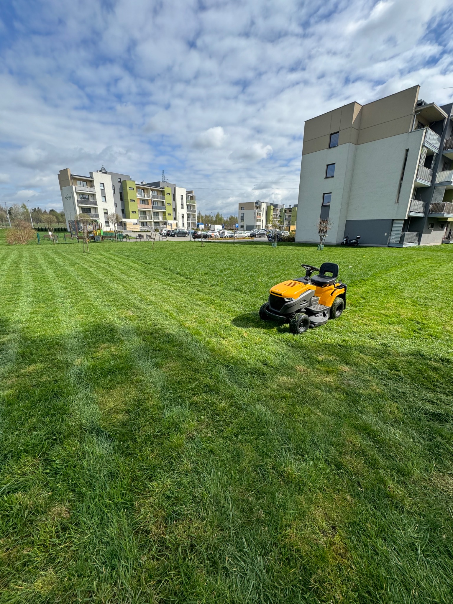 Żółta kosiarka samojezdna na tle świeżo skoszonego trawnika z widocznymi pasami, w tle budynki mieszkalne i błękitne niebo z chmurami.