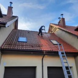 Pracownik w kombinezonie roboczym czyści czerwoną dachówkę z wykorzystaniem myjki ciśnieniowej. Część dachu jest już umyta, ukazując różnicę w kolorze. Aluminiowa drabina oparta o ścianę budynku...