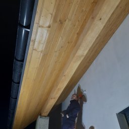 Usługi Wysokościowe, Budowlane i Szkoleniowe EIGER- Piotr Pleszczuk - Pracownik na drabinie maluje fragment podbitki dachowej na zewnątrz budynku, widoczne rynny i betonowa podpora.