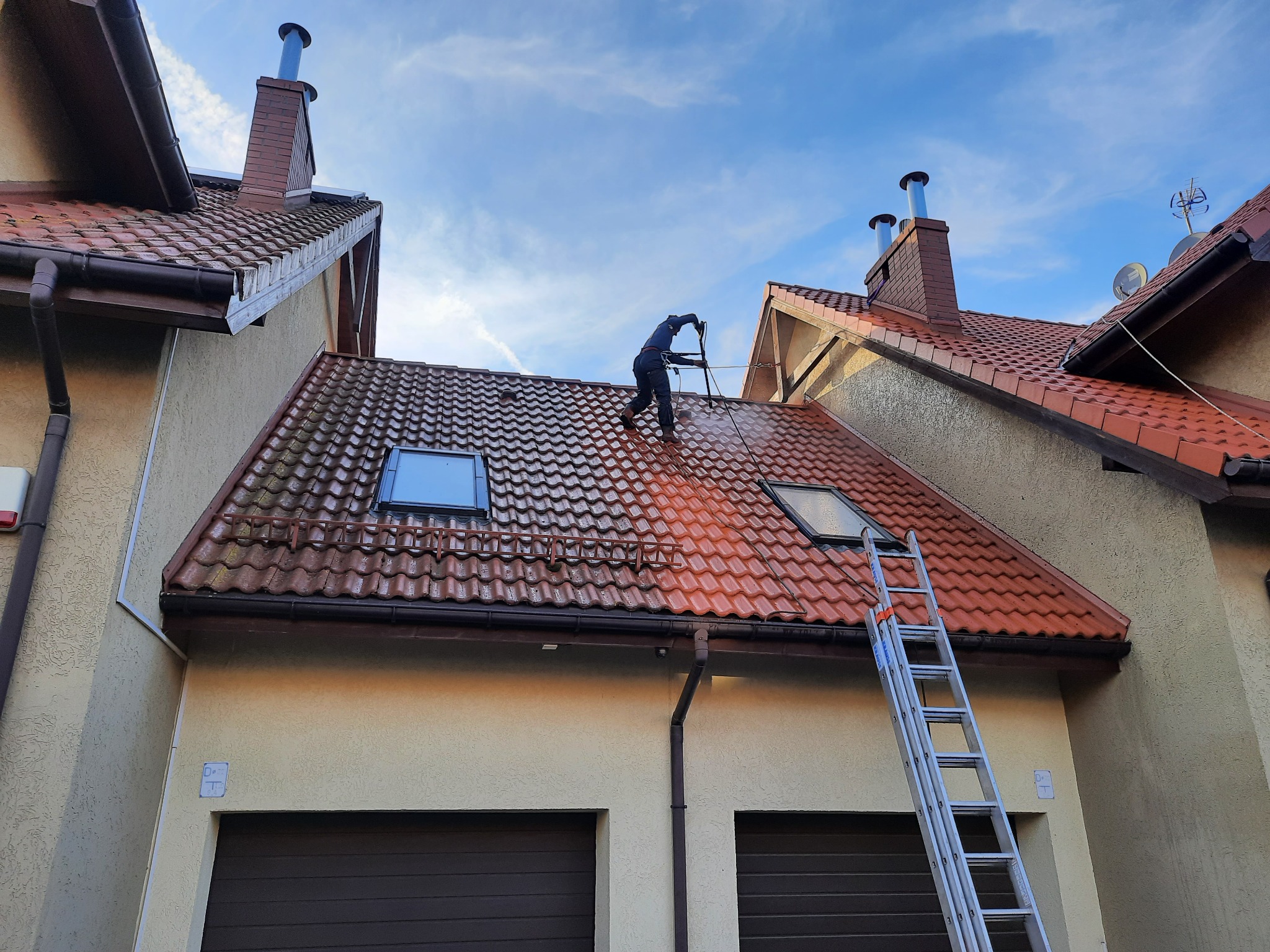 Pracownik w kombinezonie roboczym czyści czerwoną dachówkę z wykorzystaniem myjki ciśnieniowej. Część dachu jest już umyta, ukazując różnicę w kolorze. Aluminiowa drabina oparta o ścianę budynku...