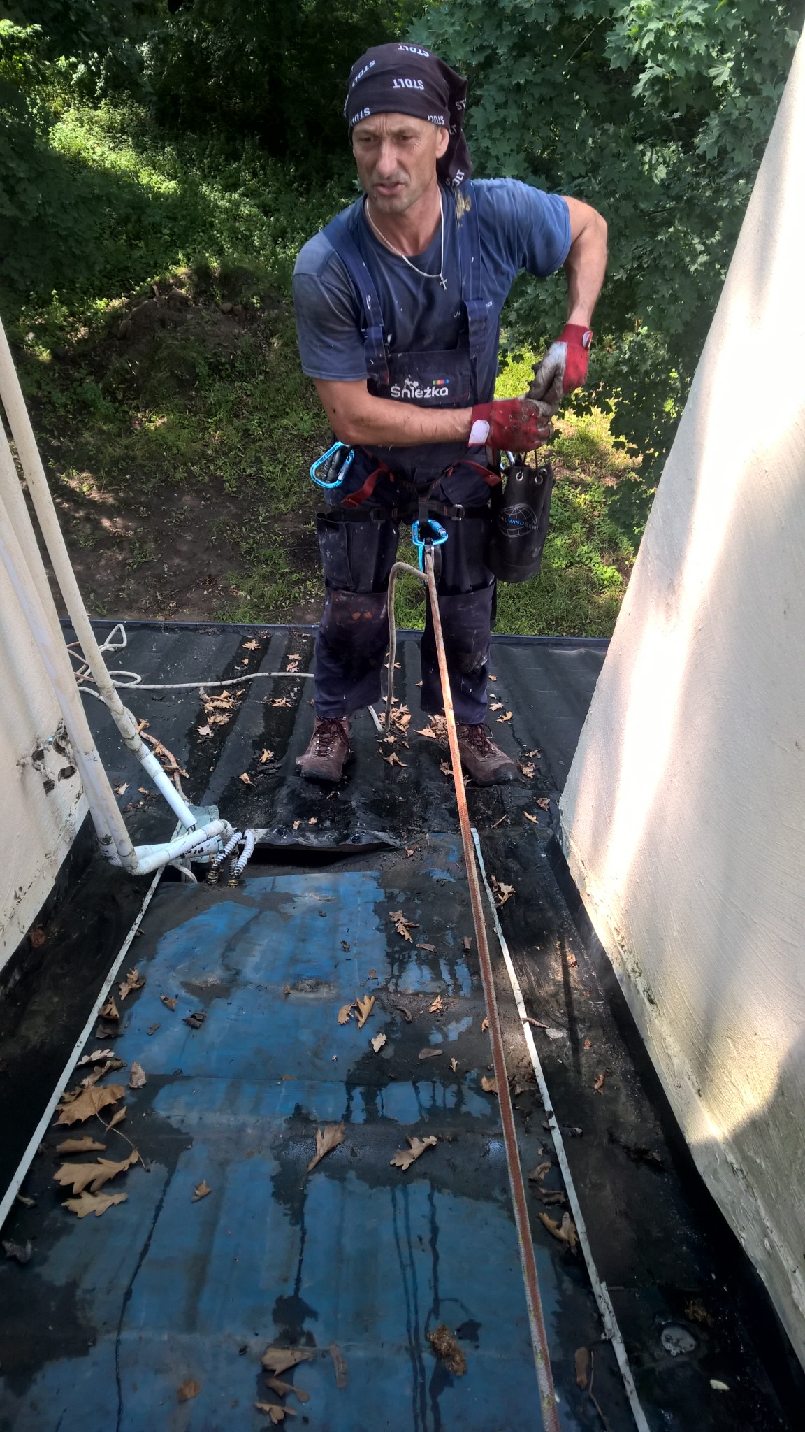 Pracownik w kombinezonie firmy Śnieżka, zabezpieczony linami, na mokrym dachu z liśćmi, podczas pracy wysokościowej.