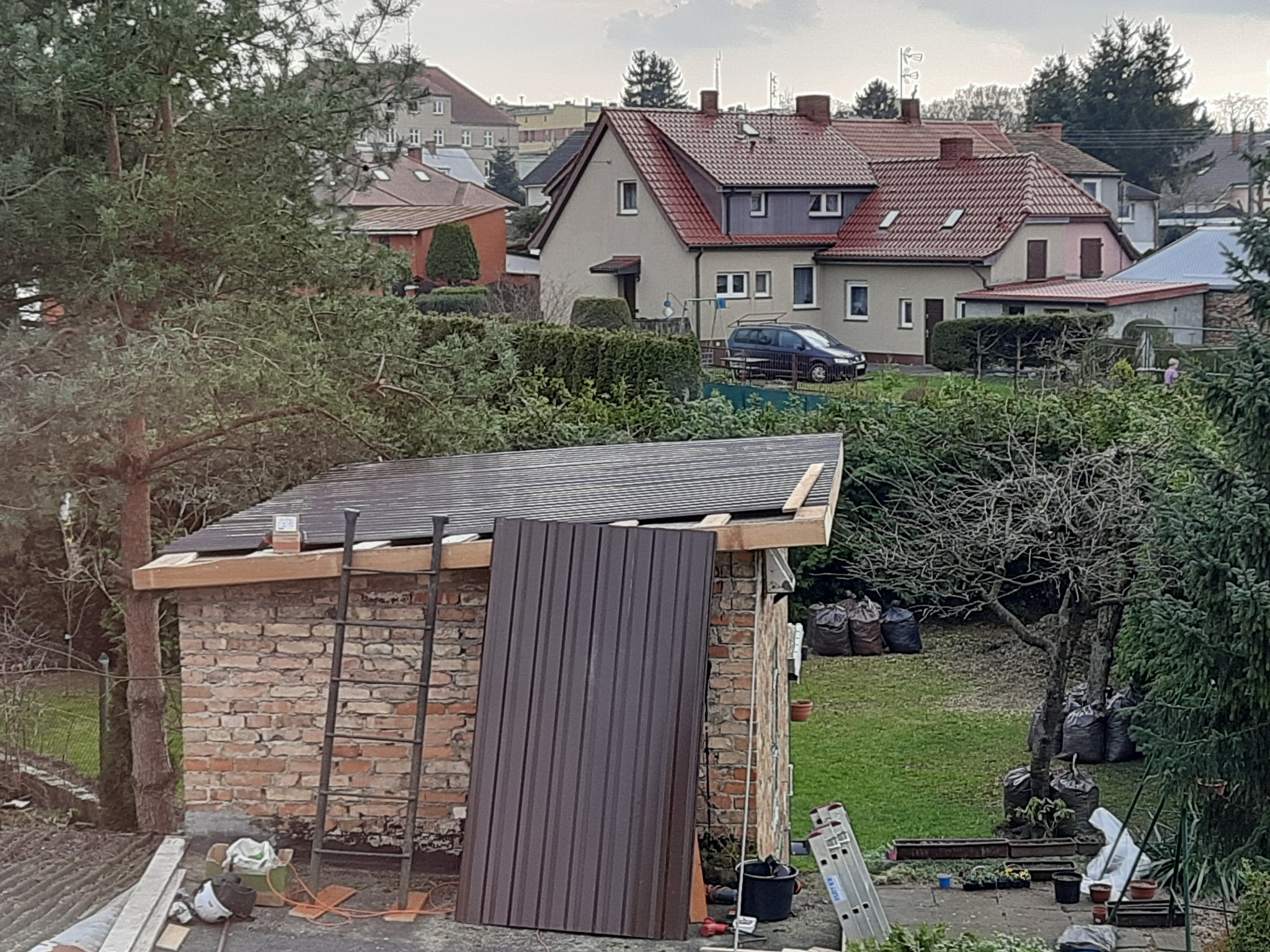 Budowa dachu na murowanym budynku gospodarczym z użyciem metalowej blachy trapezowej; w tle domy jednorodzinne i drzewa.