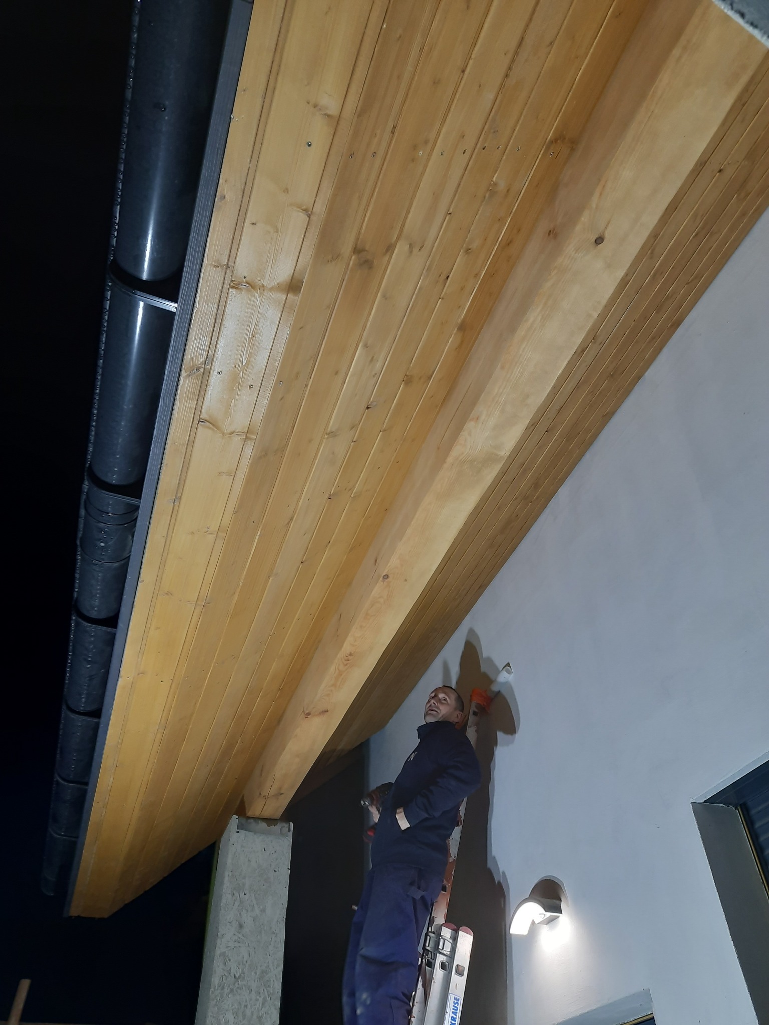 Pracownik na drabinie maluje fragment podbitki dachowej na zewnątrz budynku, widoczne rynny i betonowa podpora.