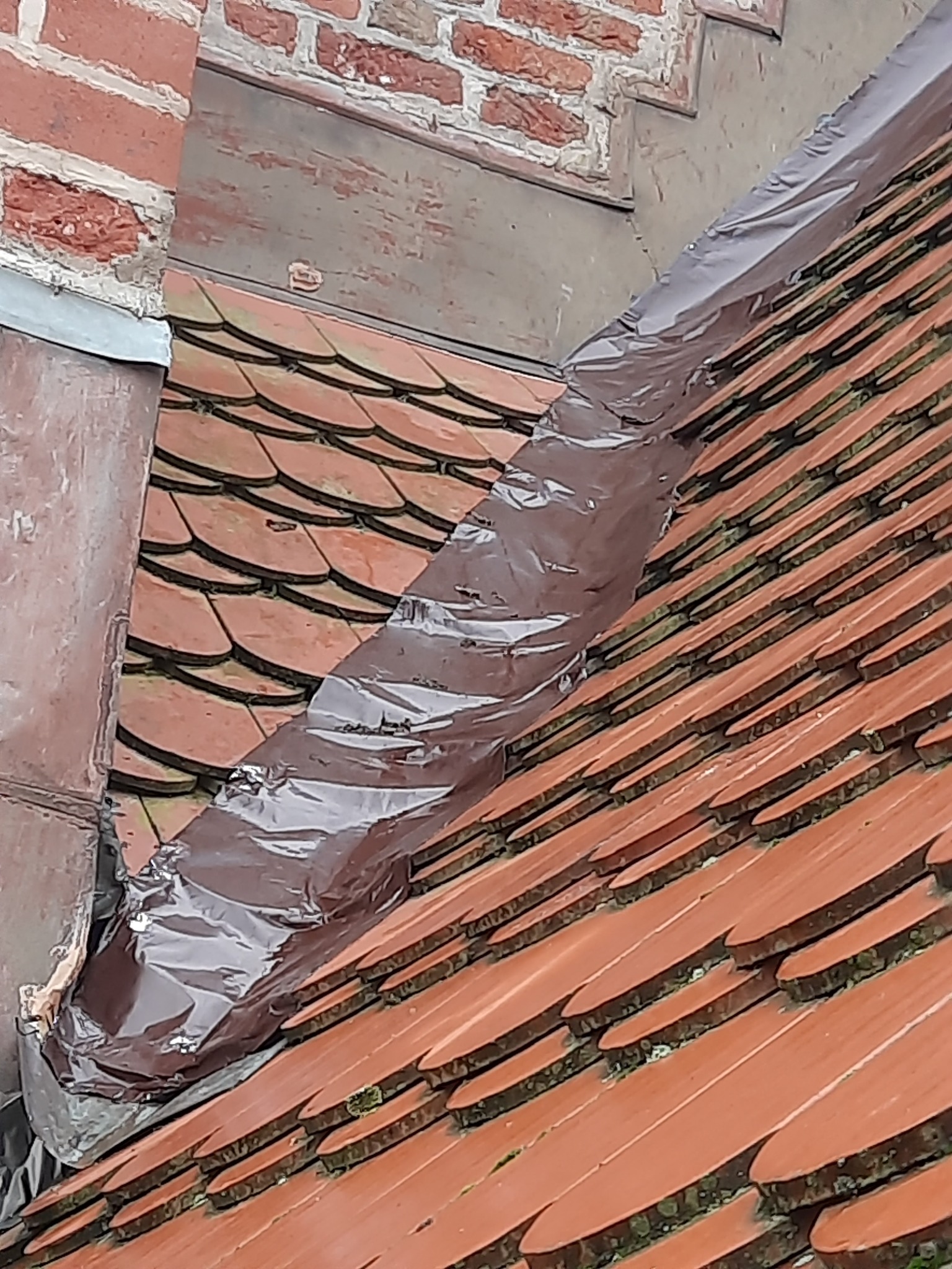 Fragment dachu krytego czerwoną dachówką, z uszczelnieniem z folii przy kominie, widoczne zabrudzenia między dachówkami.