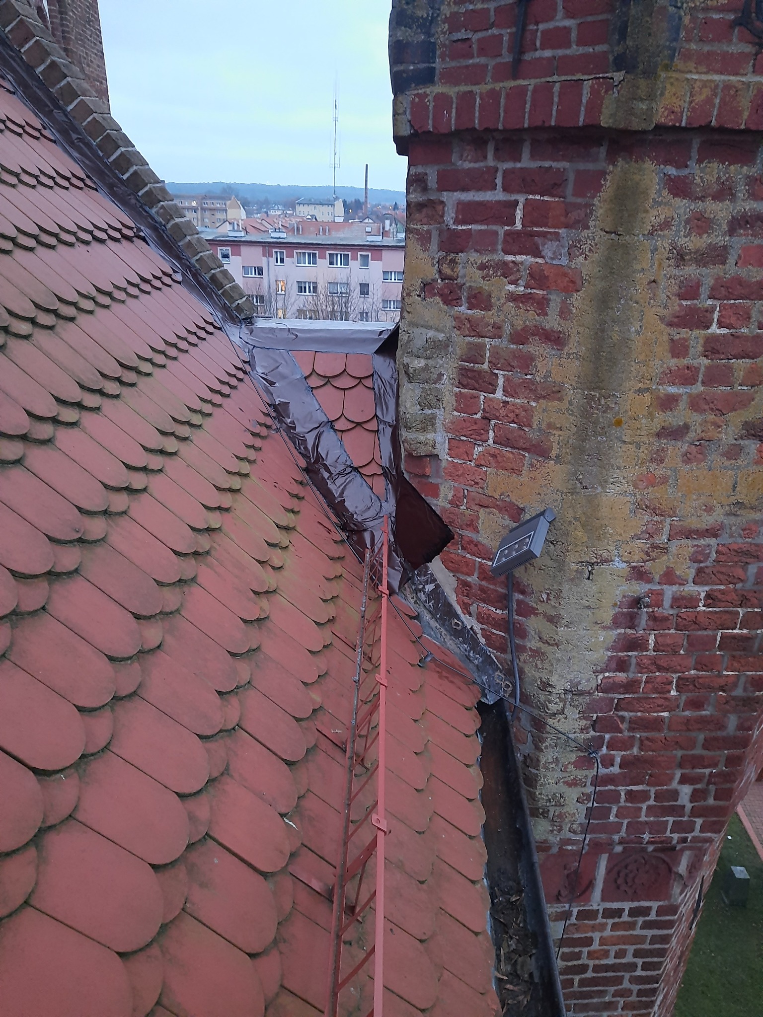 Czerwona dachówka na dachu sąsiaduje z ceglanym kominem, obok którego widać brudną rynnę wypełnioną liśćmi, a w tle zabudowania miejskie.