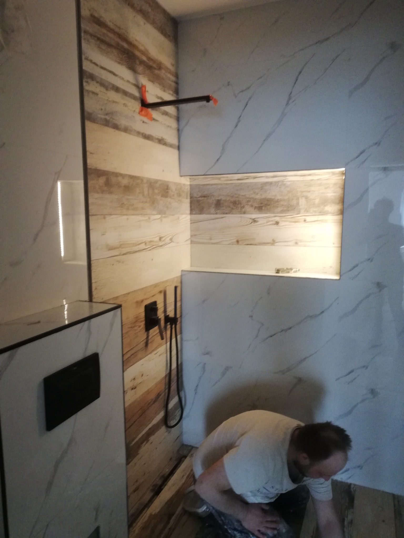 Pracownik w trakcie układania płytek podłogowych w łazience z wykończonymi ścianami w dwóch stylach: imitacja drewna i marmur z szarymi żyłami. Widoczna wnęka w ścianie z podświetleniem.