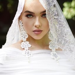 Natallia Shved - Portret kobiety z blond włosami i zielonymi oczami, ubrana w białą sukienkę i koronkowy welon z kwiatowymi kolczykami, na rozmytym zielonym tle.