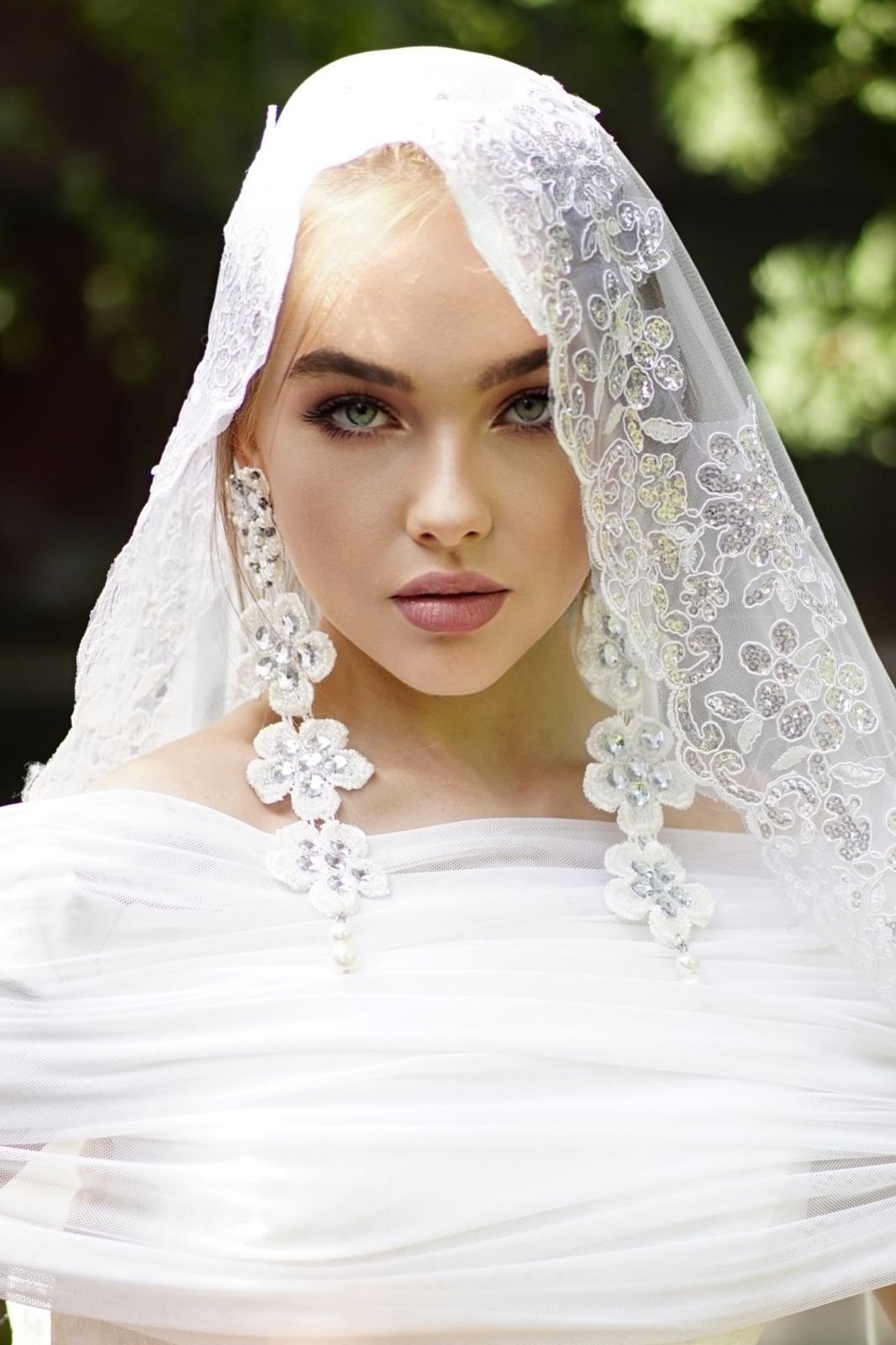 Portret kobiety z blond włosami i zielonymi oczami, ubrana w białą sukienkę i koronkowy welon z kwiatowymi kolczykami, na rozmytym zielonym tle.
