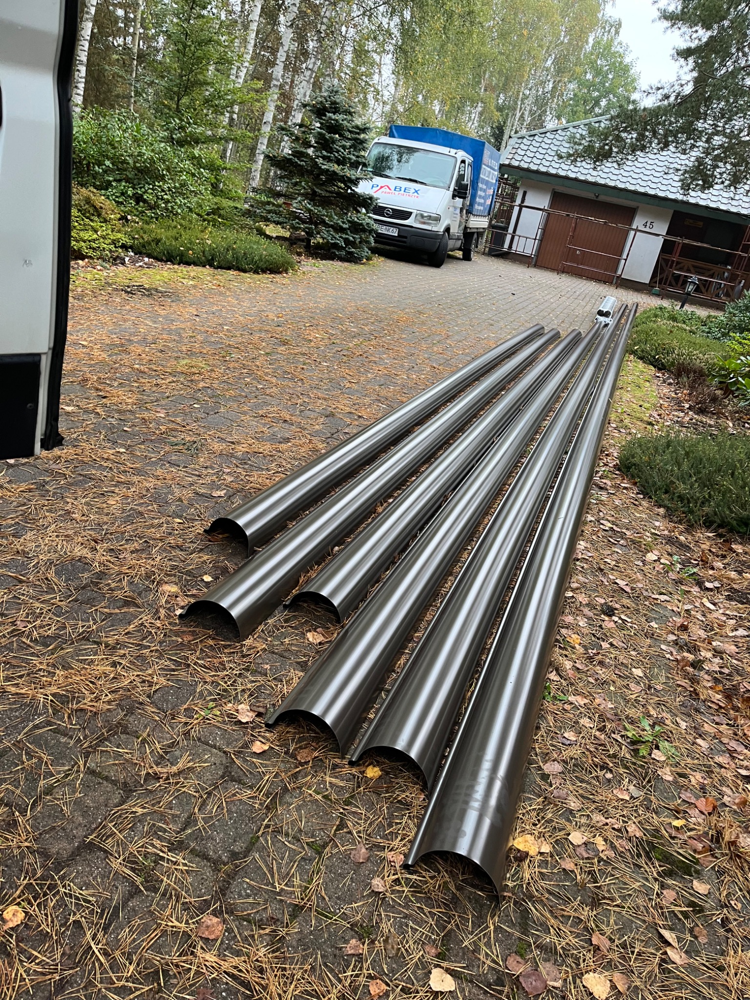 Stos ciemnych, metalowych rynien leżących na brukowanym podjeździe, z widocznym samochodem dostawczym firmy Abex i domem w tle.