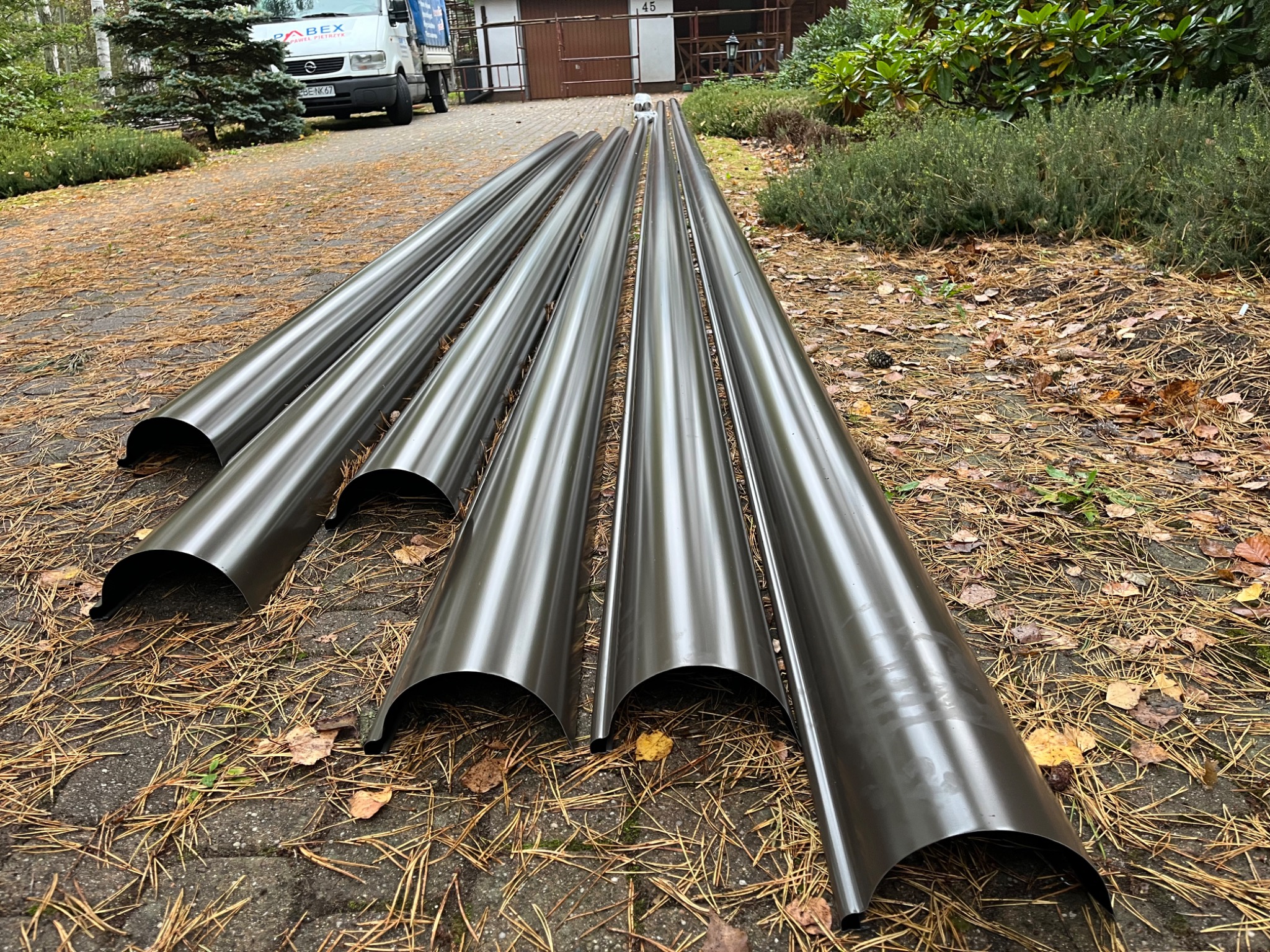 Ciemne, metalowe elementy systemu odprowadzania wody deszczowej leżące na brukowanej powierzchni z igliwiem i liśćmi, gotowe do montażu.