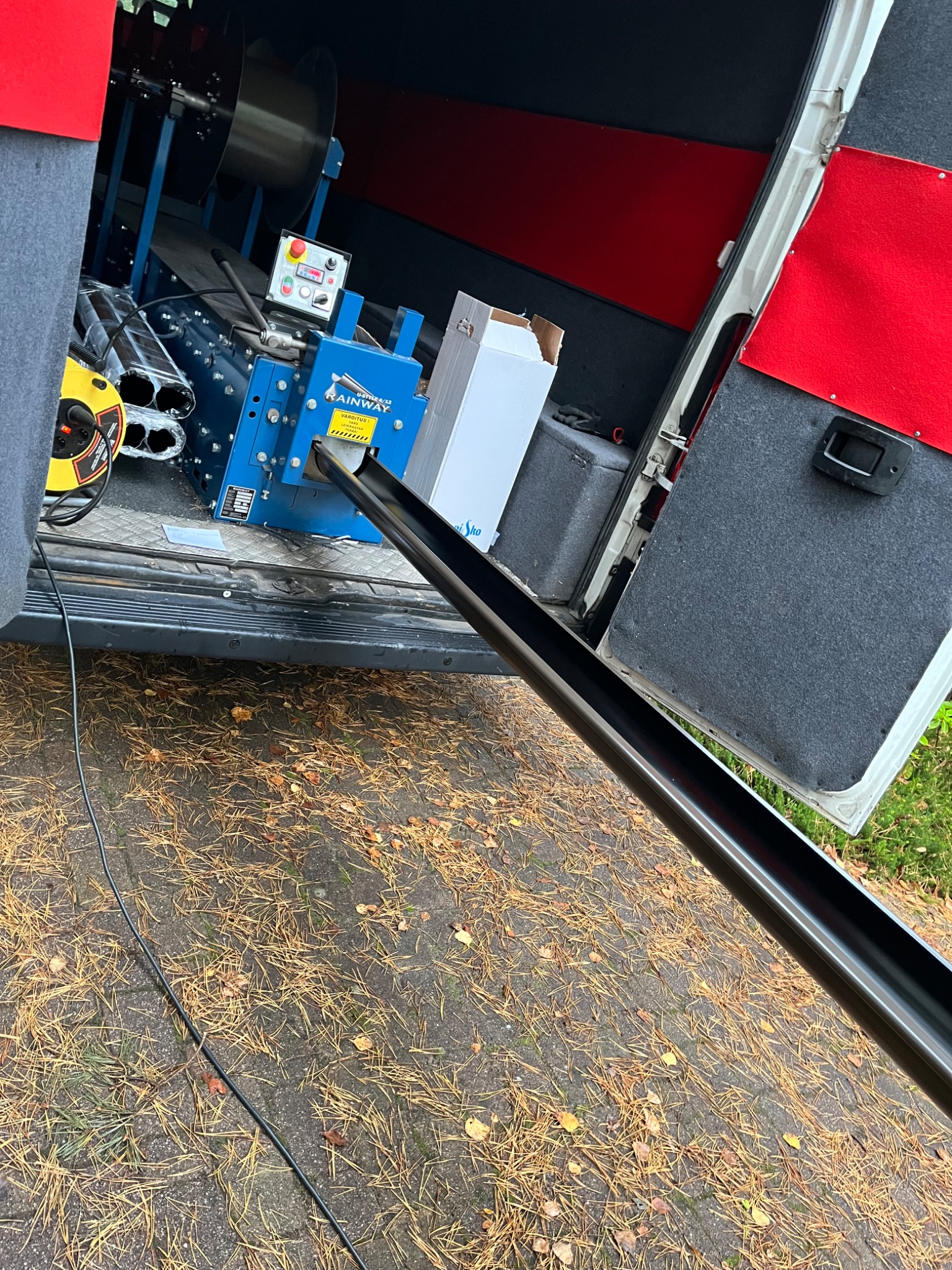 Wnętrze samochodu dostawczego z maszyną do formowania elementów, widoczny profilowany element w kolorze czarnym wychodzący na zewnątrz, na ziemi igliwie.