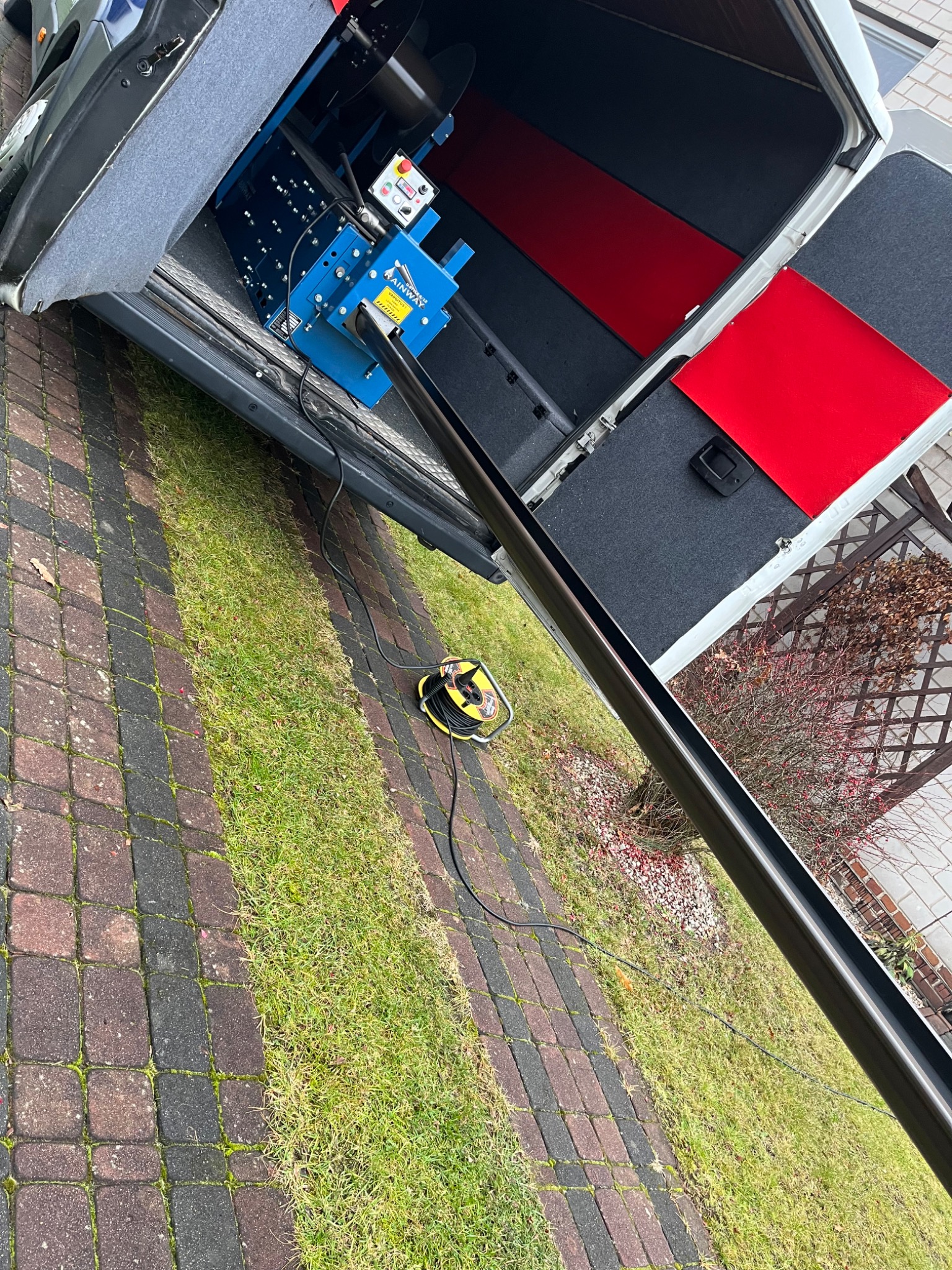 Mobilna giętarka do rynien Rainway w bagażniku busa, z widoczną długą rynną wysuwaną w kierunku trawnika i kostki brukowej.