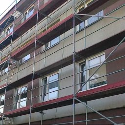 Max Dach - Budynek wielorodzinny w trakcie prac elewacyjnych. Widoczne rusztowania i fragmenty ocieplonej oraz pomalowanej ściany w odcieniach szarości i brązu.