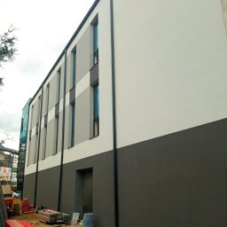 Max Dach - Nowoczesna elewacja budynku po remoncie, z wyraźnym podziałem na szary dół i białą górę. Widoczne okna i rynny. Na dole materiały budowlane.