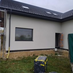 Max Dach - Jasna elewacja domu jednorodzinnego z czarną dachówką i oknami. Na pierwszym planie wiadro z farbą i narzędzia. Częściowo widoczne rusztowanie.