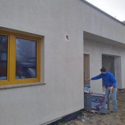 Max Dach - Pracownik w trakcie malowania elewacji domu jednorodzinnego na szaro. Widoczne okno z żółtą ramą, puszka z farbą i narzędzia. Fragment ocieplenia przy ziemi.