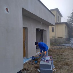 Max Dach - Pracownik w niebieskiej bluzie wygładza szarą elewację domu jednorodzinnego. Widoczne bloczki, narzędzia i fragmenty ocieplenia. Mglista pogoda w tle.