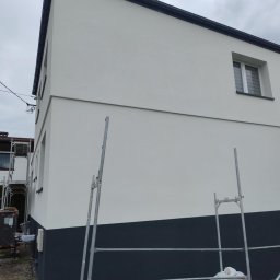 Max Dach - Dwukolorowa elewacja domu po malowaniu, z widocznymi fragmentami rusztowania. Górna część ściany biała, dolna ciemnoszara. Okno z białymi ramami. Pochmurne niebo w tle.